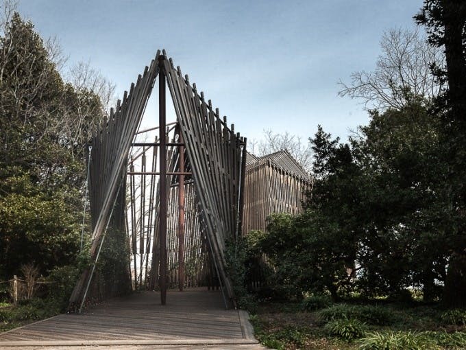 Деревянная конструкция с треугольным входом, окруженная деревьями и зеленью под чистым небом. Путь, ведущий внутрь.