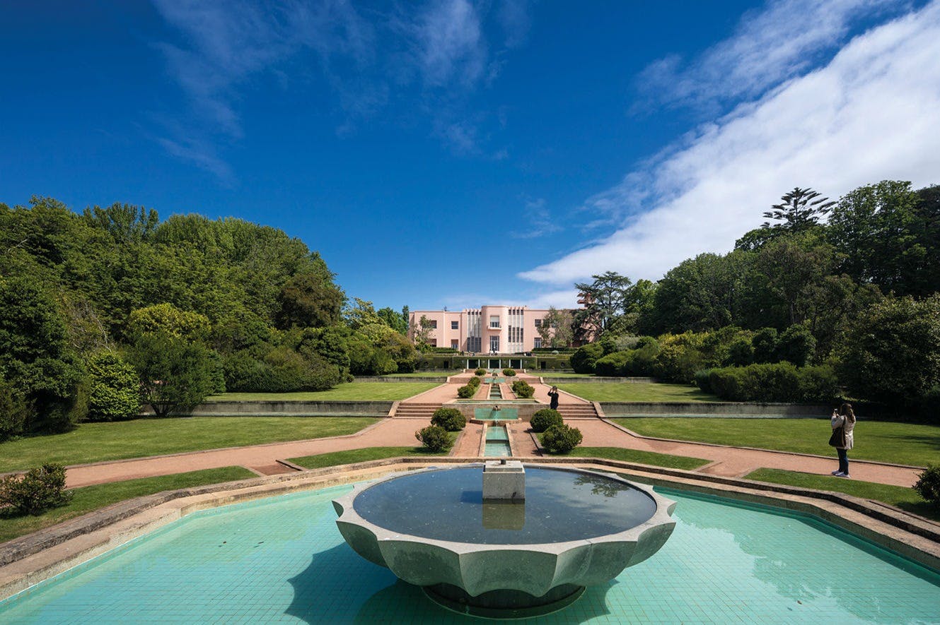 푸른 하늘 아래 녹음으로 둘러싸인 크고 분홍색 건물로 이어지는 중앙 분수가 있는 정원이 있습니다.