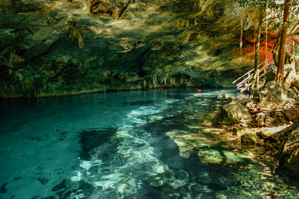 Clear blue water in an underground cave, with rock formations and two swimmers in the background. Sunlight illuminates the water.