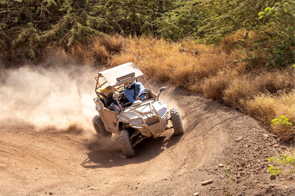 Two people driving a dune buggy on a dirt path surrounded by dry vegetation, with dust trailing behind the vehicle.