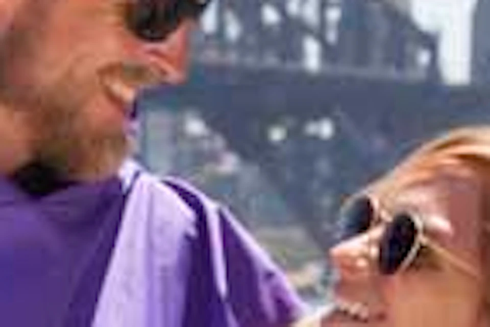A couple wearing purple ponchos smiles at each other near water with the Sydney Harbour Bridge visible in the background.
