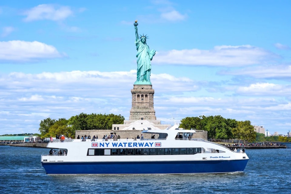 A ferry labeled 'NY Waterway' passes in front of the Statue of Liberty on a sunny day with scattered clouds.