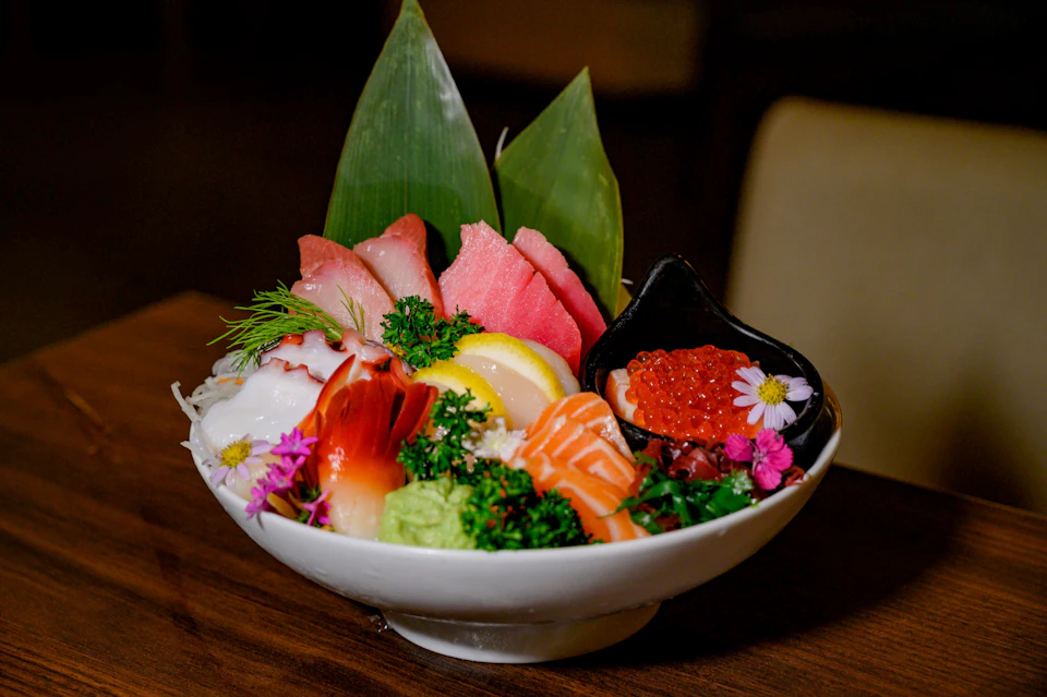 A bowl of assorted sashimi, garnished with flowers and greens, featuring salmon, tuna, roe, and lemon slices.