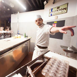 En kock i vit uniform ger en demonstration till en grupp barn som står bakom en disk, med chokladformar i närheten.