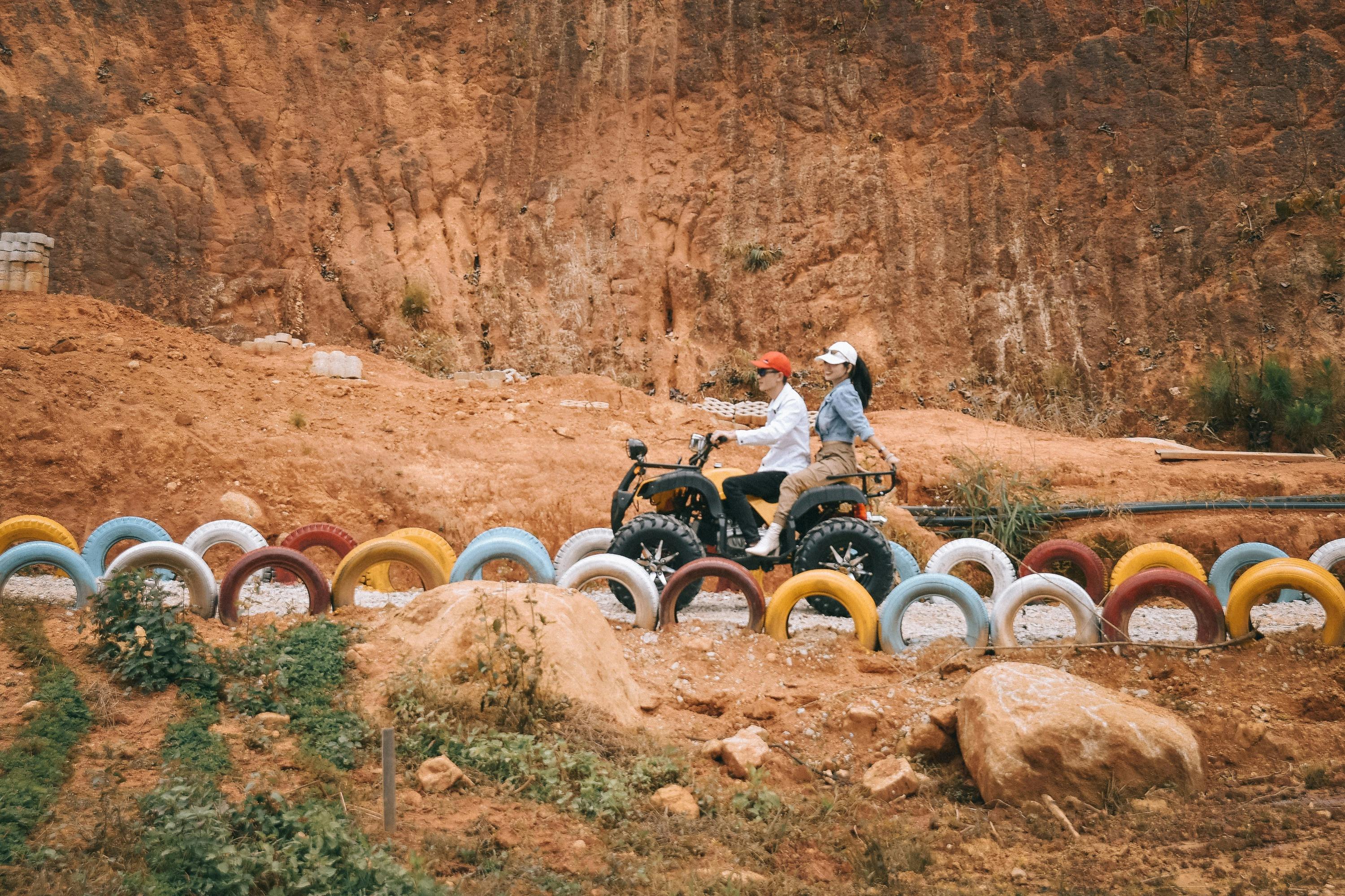 Two people riding a quad bike across a dirt track lined with colorful, half-buried tires, with a rocky hill in the background.
