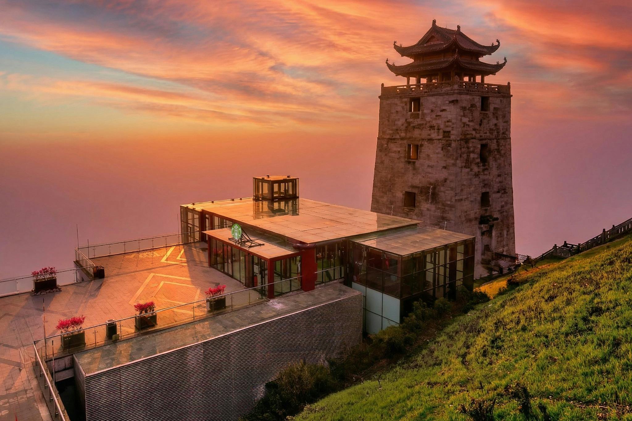 A traditional Chinese-style tower next to a modern building at sunset, situated on a grassy hillside.