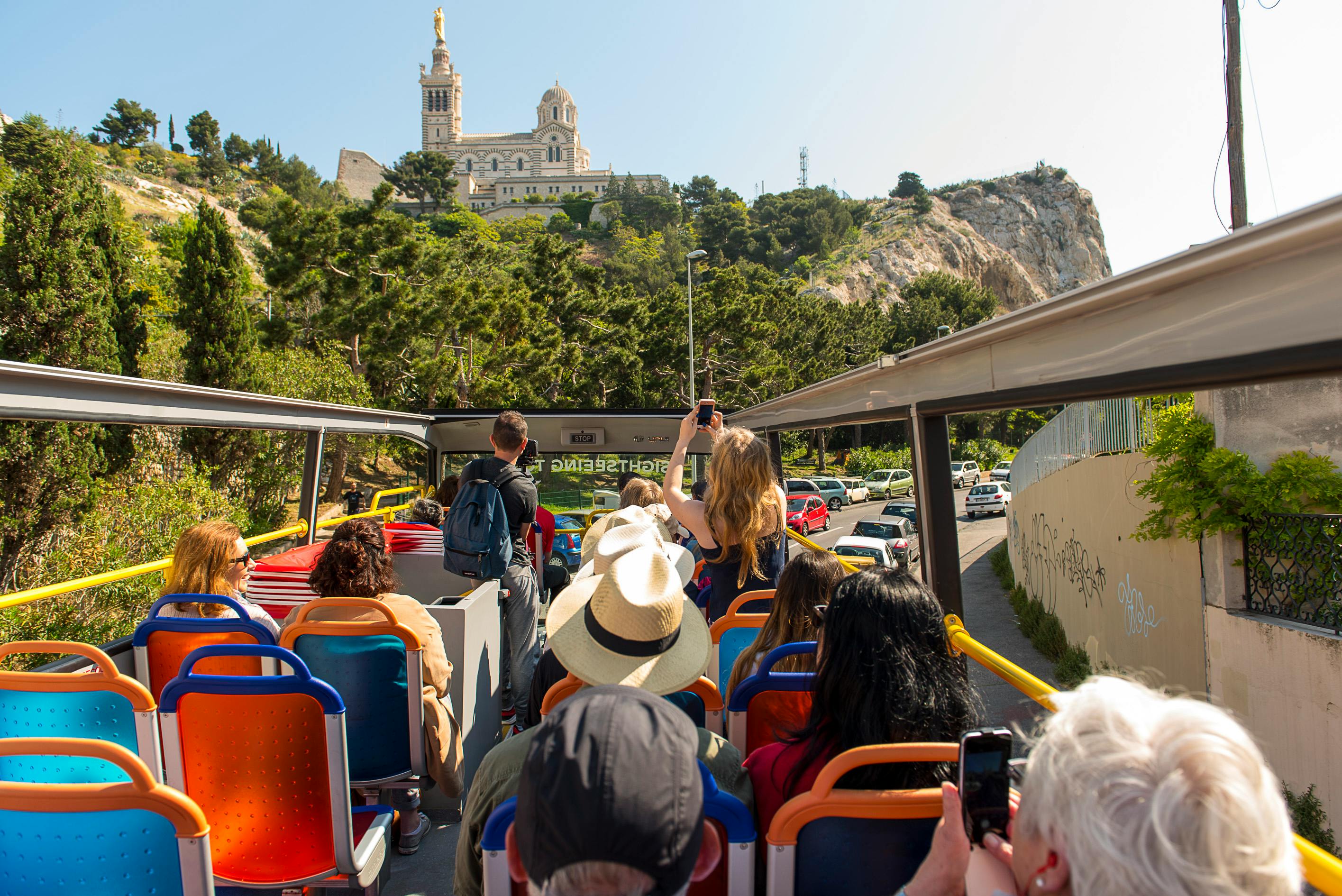 Turister på en öppen buss tar bilder av en kyrka på en kulle omgiven av träd och en väg med parkerade bilar.