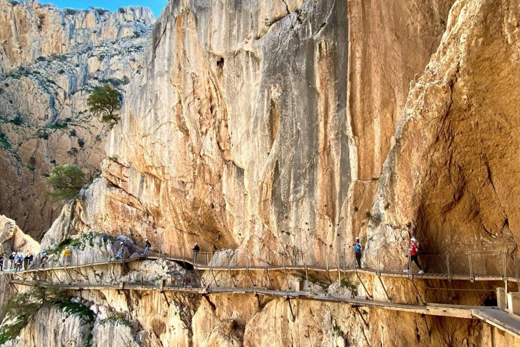 People walking on a narrow wooden walkway attached to a steep, rocky cliff face under a clear sky.