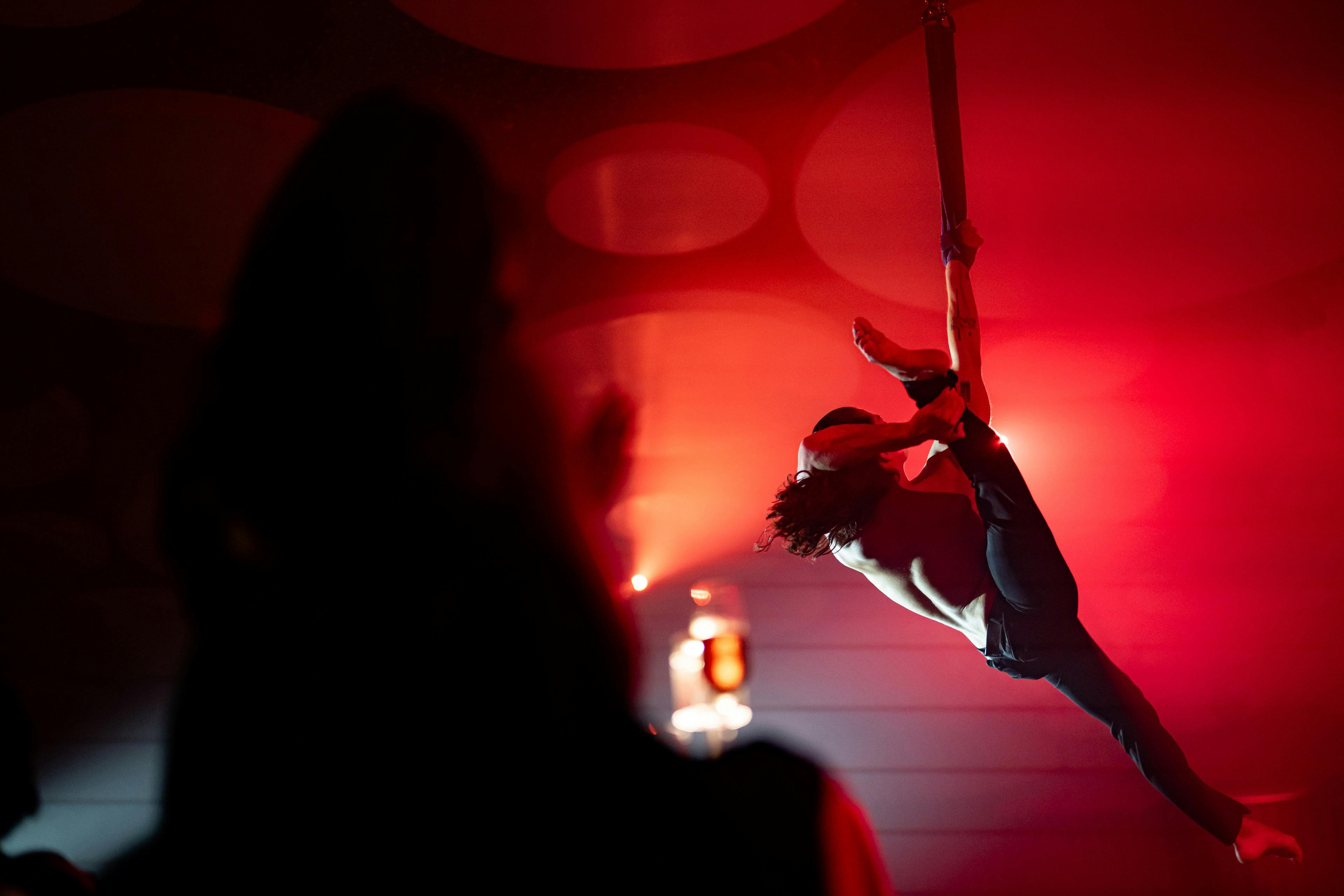 Ein Performer, der an einem Gurt in der Luft hängt, unter roter Beleuchtung, mit einem Zuschauer als Silhouette im Vordergrund.