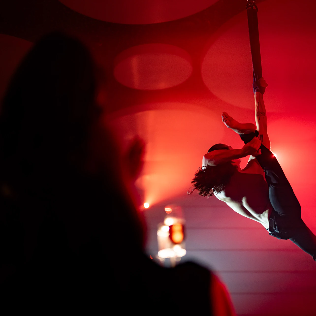 A performer suspended by a strap in mid-air under red lighting, with an audience member silhouetted in the foreground.