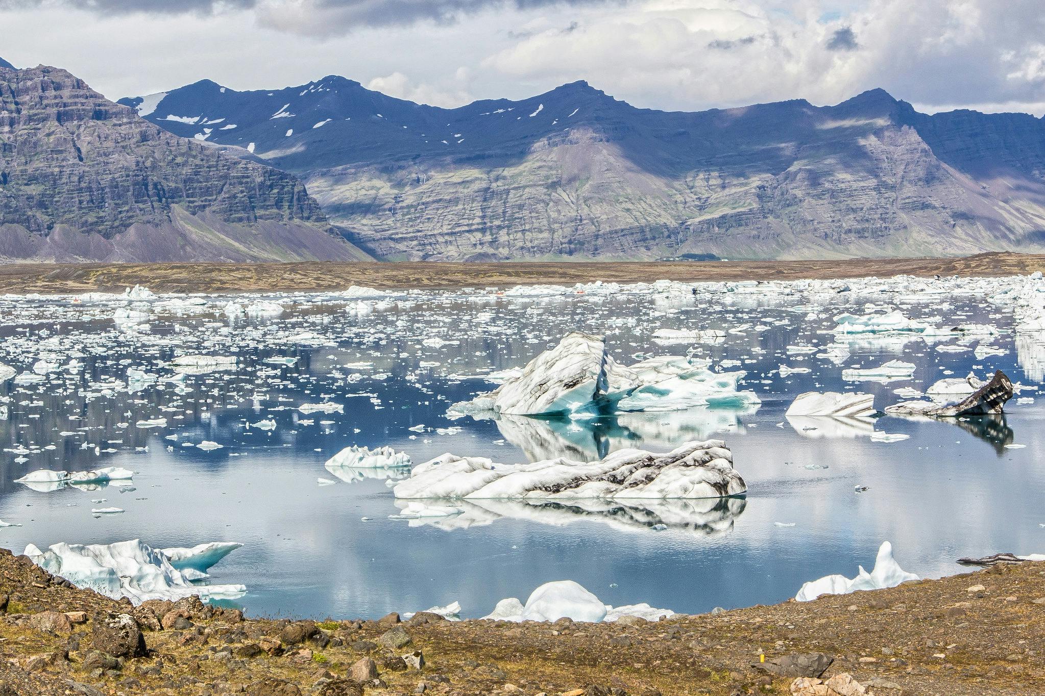 Icebergs flottants sur un lac calme et réfléchissant, entouré de rochers et de montagnes sous un ciel nuageux.