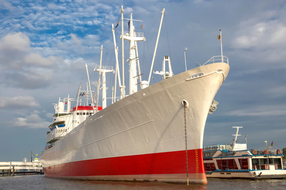 Large white and red ship docked by the water with a blue sky and clouds in the background.