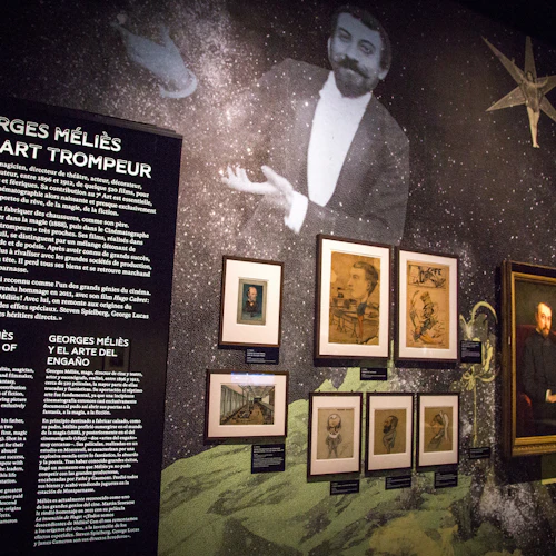 Utställ vägg med text om Georges Méliès på franska, engelska och spanska, omgiven av olika inramade porträtt och illustrationer.