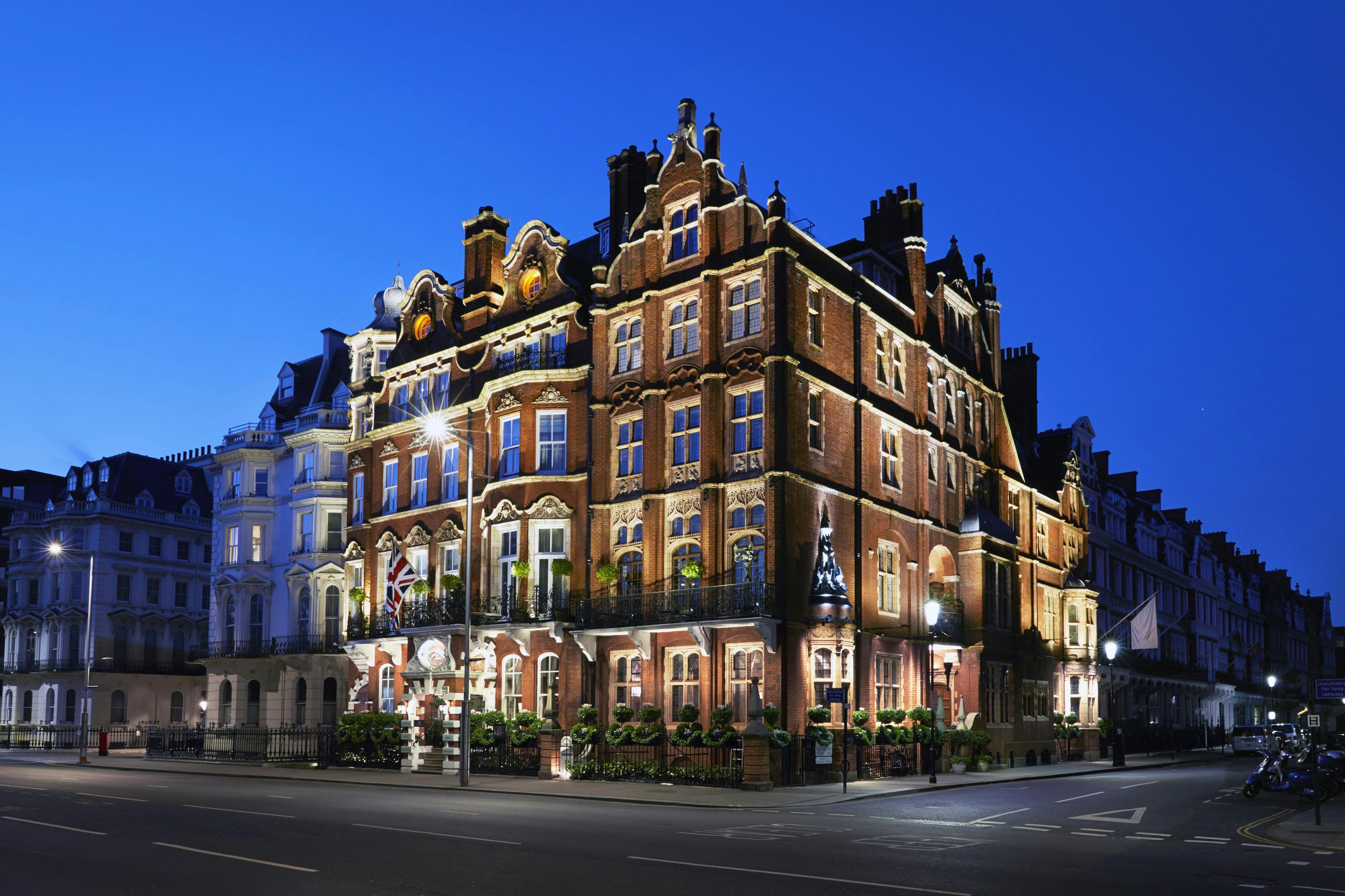 A lit-up, ornate red-brick building with decorative windows and balconies, set in an urban area under a deep blue evening sky.