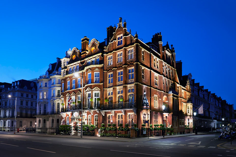 A lit-up, ornate red-brick building with decorative windows and balconies, set in an urban area under a deep blue evening sky.