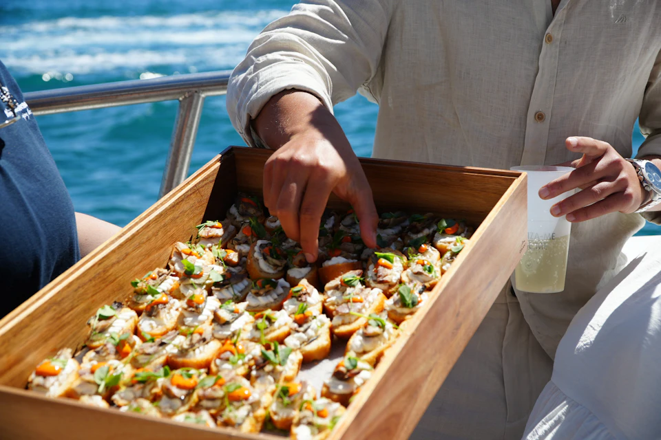 A person takes an appetizer from a wooden tray filled with finger foods, with a background of a railing and ocean waves.