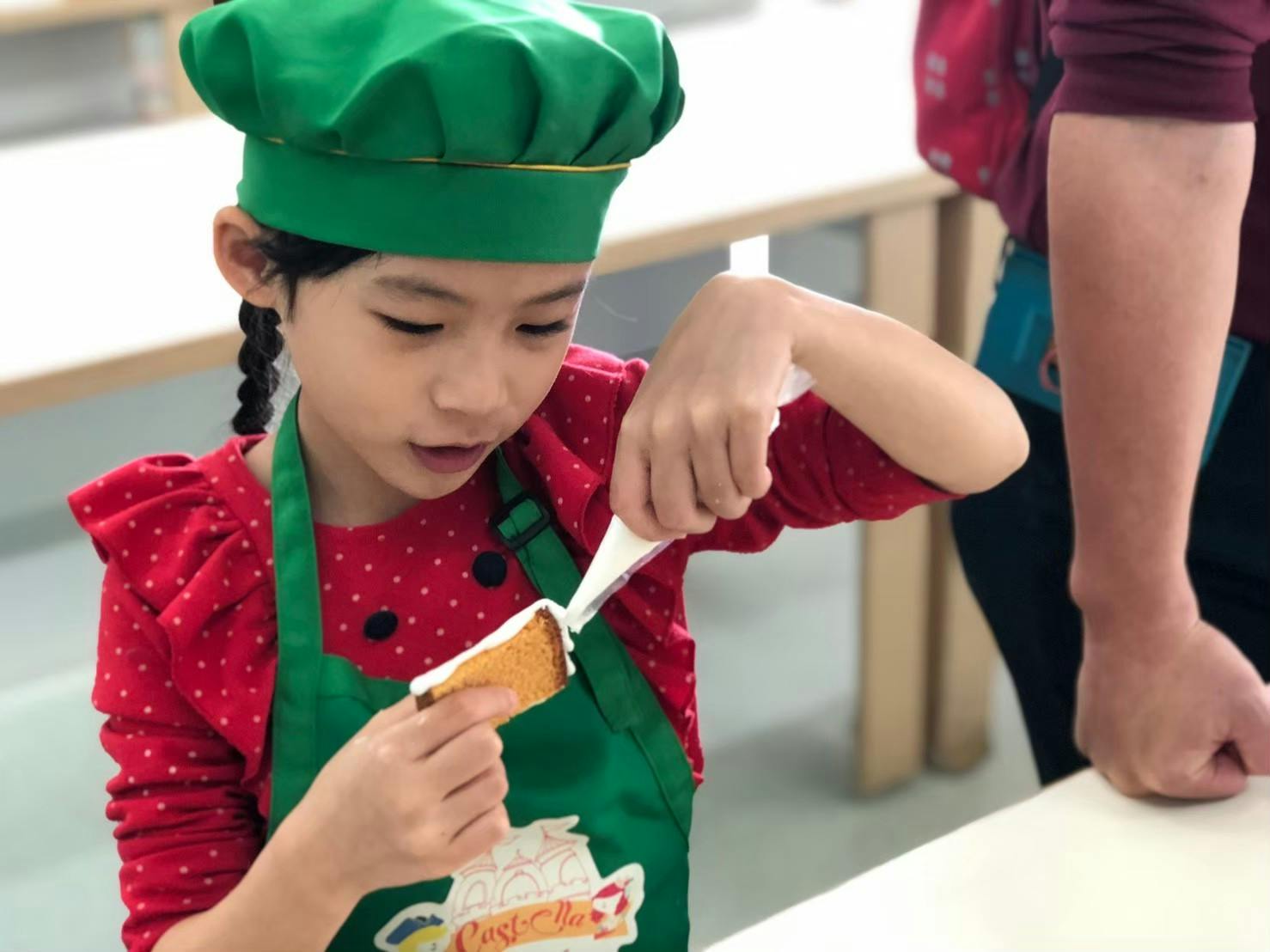 A child in a green chef hat and apron decorates a piece of cake with white icing in a classroom setting.