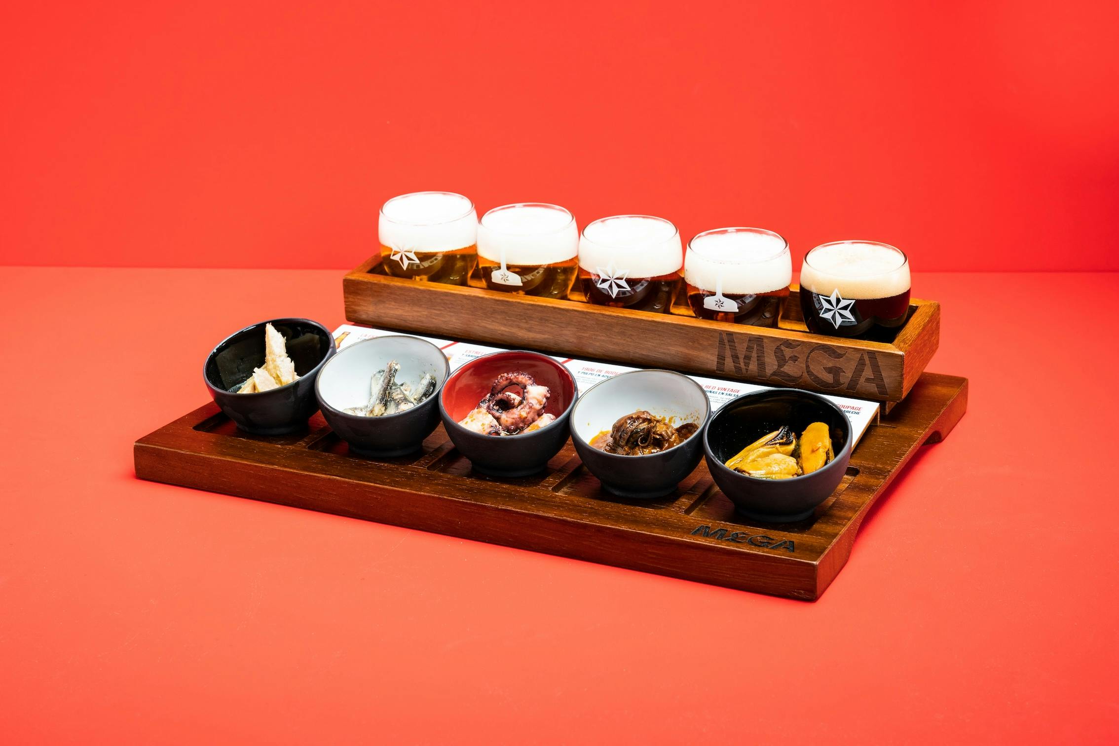 Sześć małych dań z różnymi przystawkami i cztery napełnione szklanki piwa na drewnianej tacy na czerwonym tle.