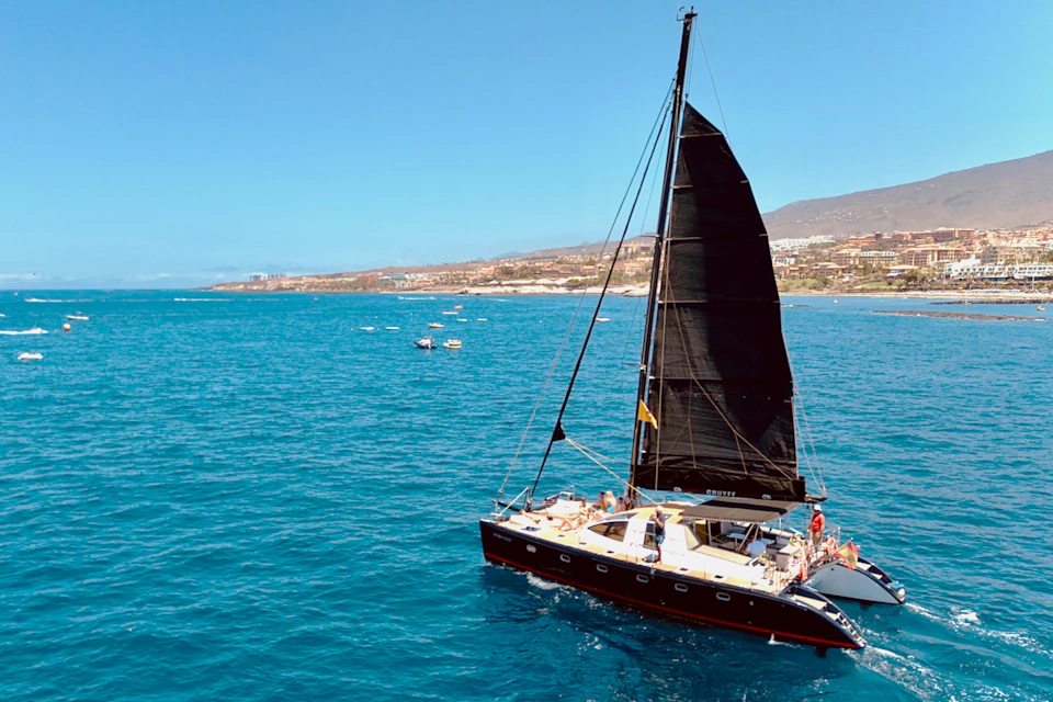 A sailboat with a black sail on a clear blue sea, with a coastline visible in the background under a sunny sky.