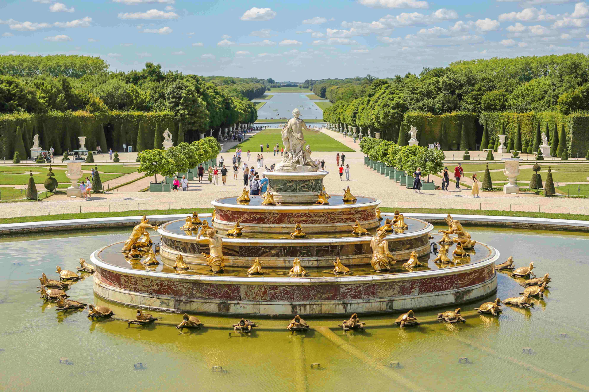 Grand fountain with golden statues in a garden with a tree-lined path and people walking under a blue sky with clouds.