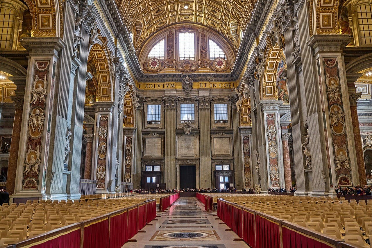Ornate interior of St. Peter's Basilica featuring high arches, red-bannered seating, detailed columns, and a gold-tiled ceiling.