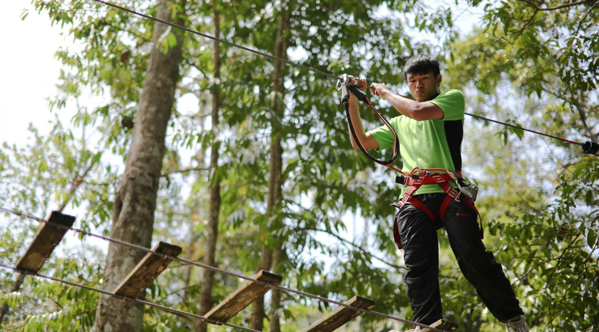 A person wearing a green shirt and safety harness is gripping a zipline cable in a forest setting.