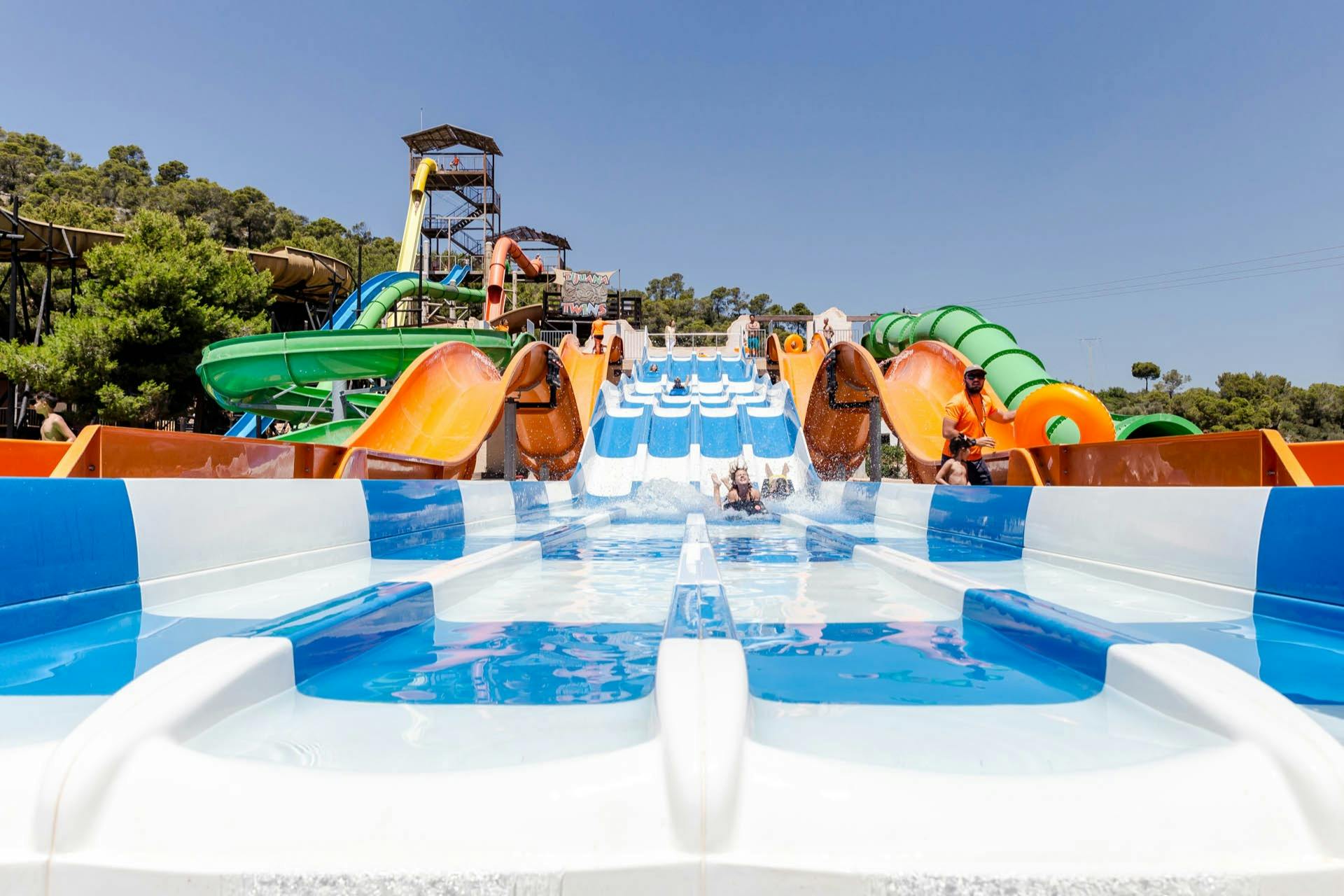 Mensen genieten van een waterglijbaan met meerdere banen in een waterpark met kleurrijke glijbanen en structuren onder een strakblauwe hemel.