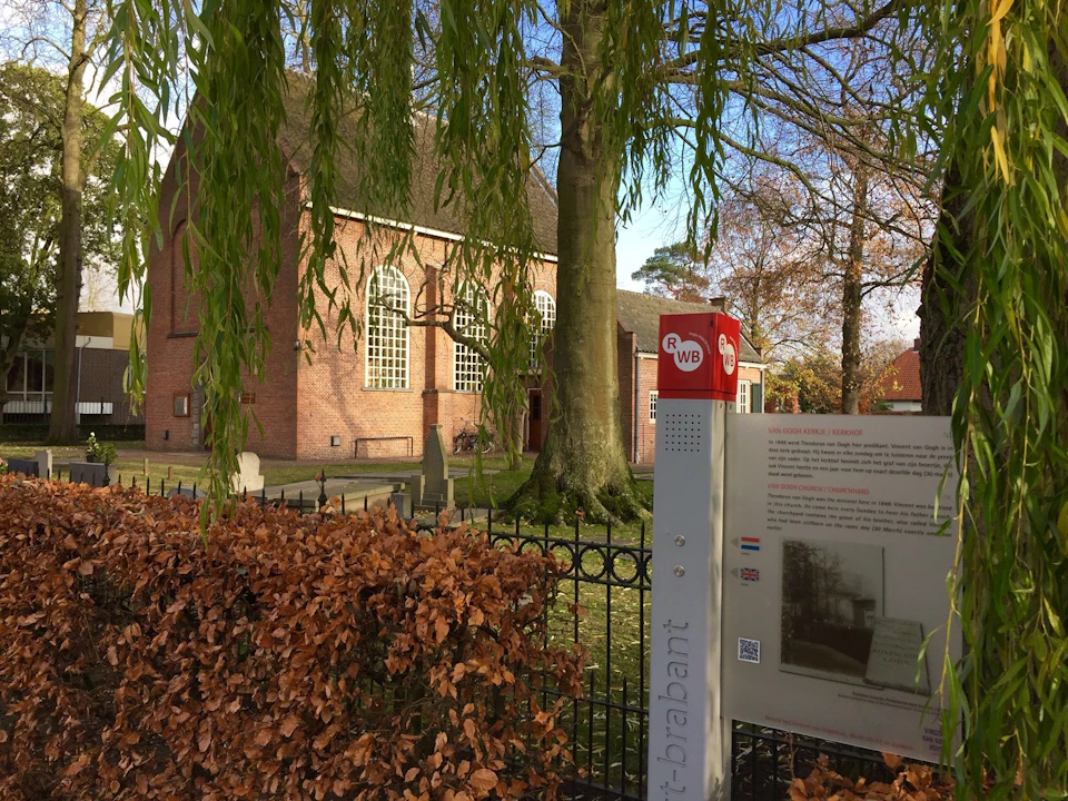 Brick building with arched windows behind trees and a fence. Informational sign and hedges in the foreground.