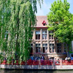 En pittoresk, historisk byggnad med röd kant och stora fönster, delvis skymd av frodiga träd, ligger bredvid en kanal. Människor sitter längs ett rött räcke.