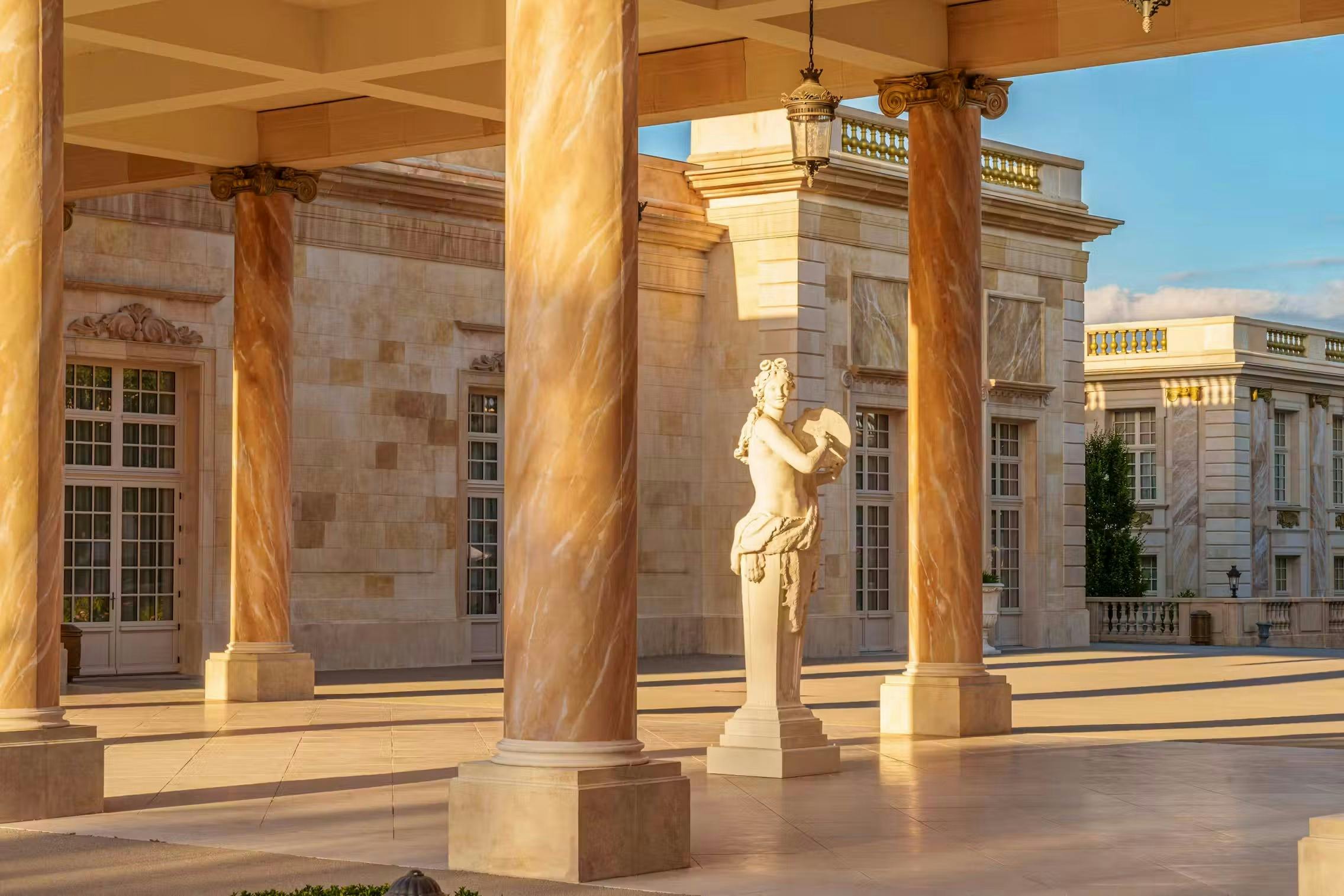 A courtyard with marble columns, a stone statue of a figure, and a classical building in the background under sunlight.