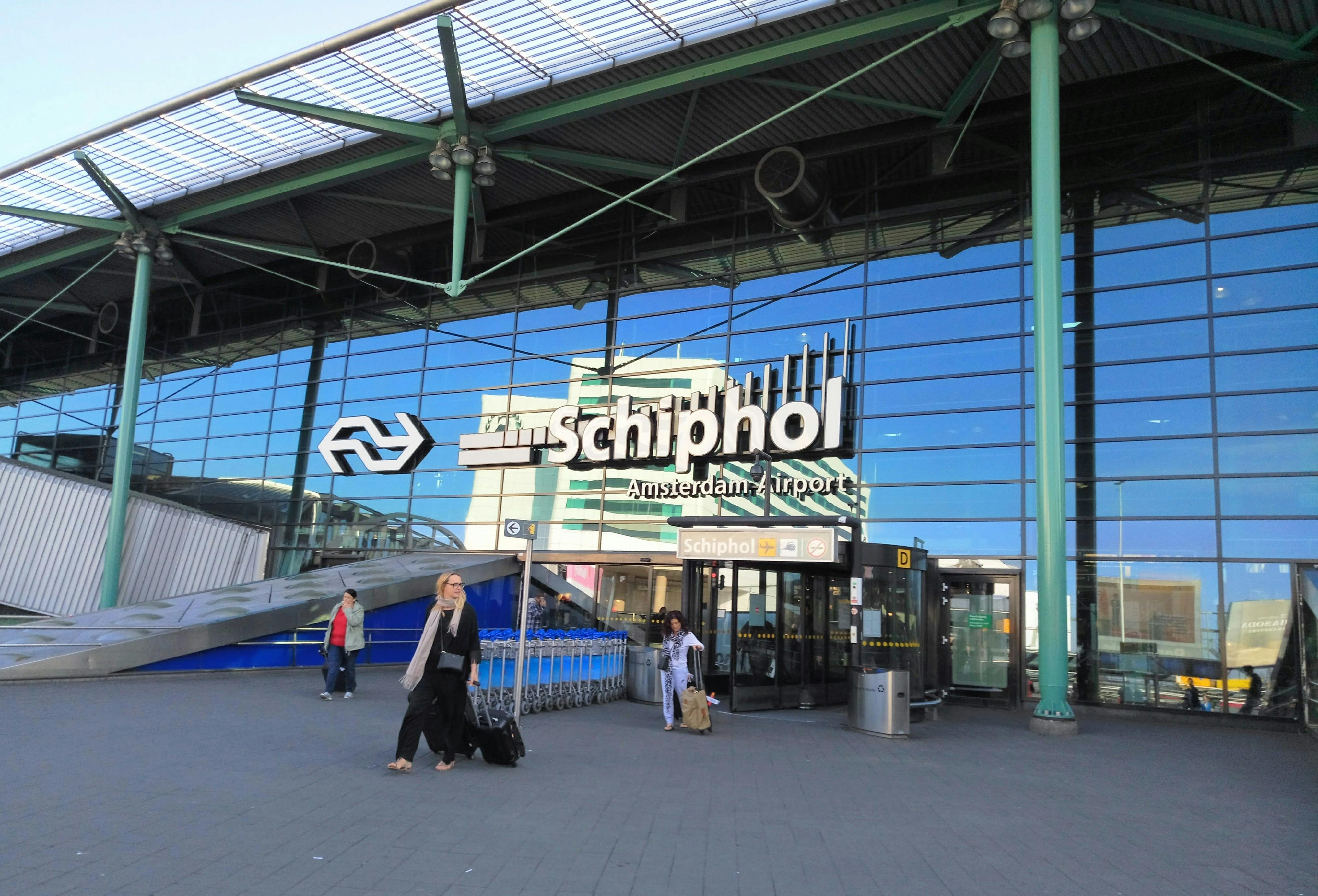 Entrada a l&#39;aeroport de Schiphol Amsterdam amb gent caminant, façana de vidre, senyalització de l&#39;aeroport i carros d&#39;equipatge.