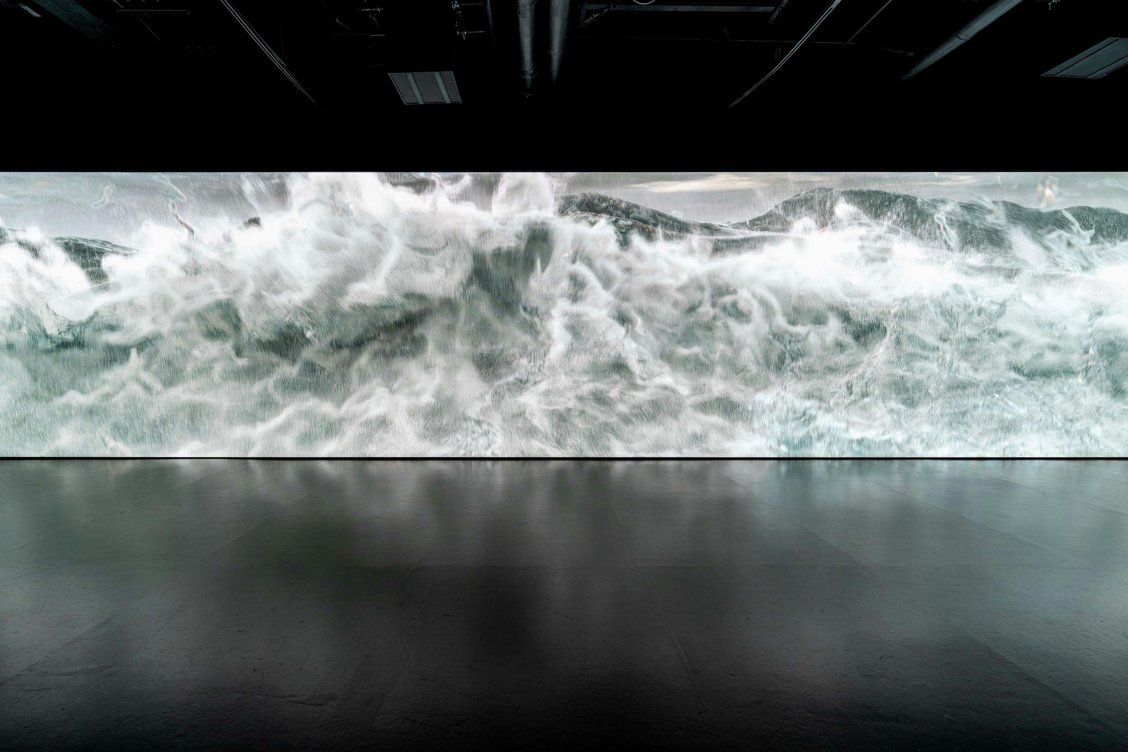 Ein großer digitaler Bildschirm zeigt ein Bild von turbulenten Wellen in einem dunklen Raum mit reflektierendem Boden.