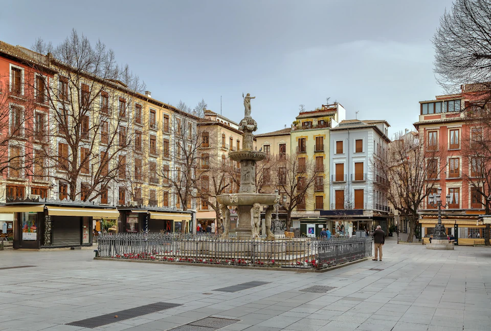 A historic stone fountain in a fenced plaza with surrounding multi-story buildings and bare trees; a couple of people are walking.