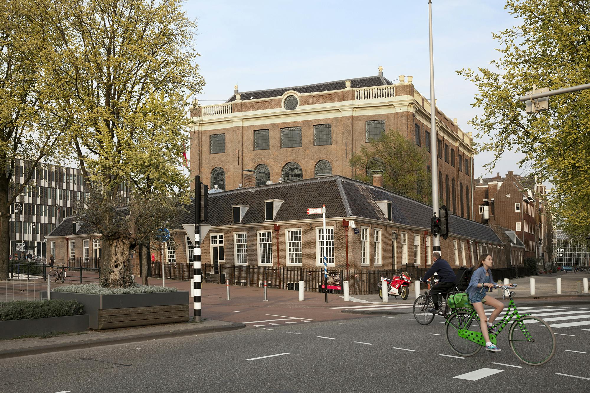 Une femme roule sur un vélo vert tandis qu'un homme marche dans une rue à côté d'un bâtiment historique en briques et d'arbres.
