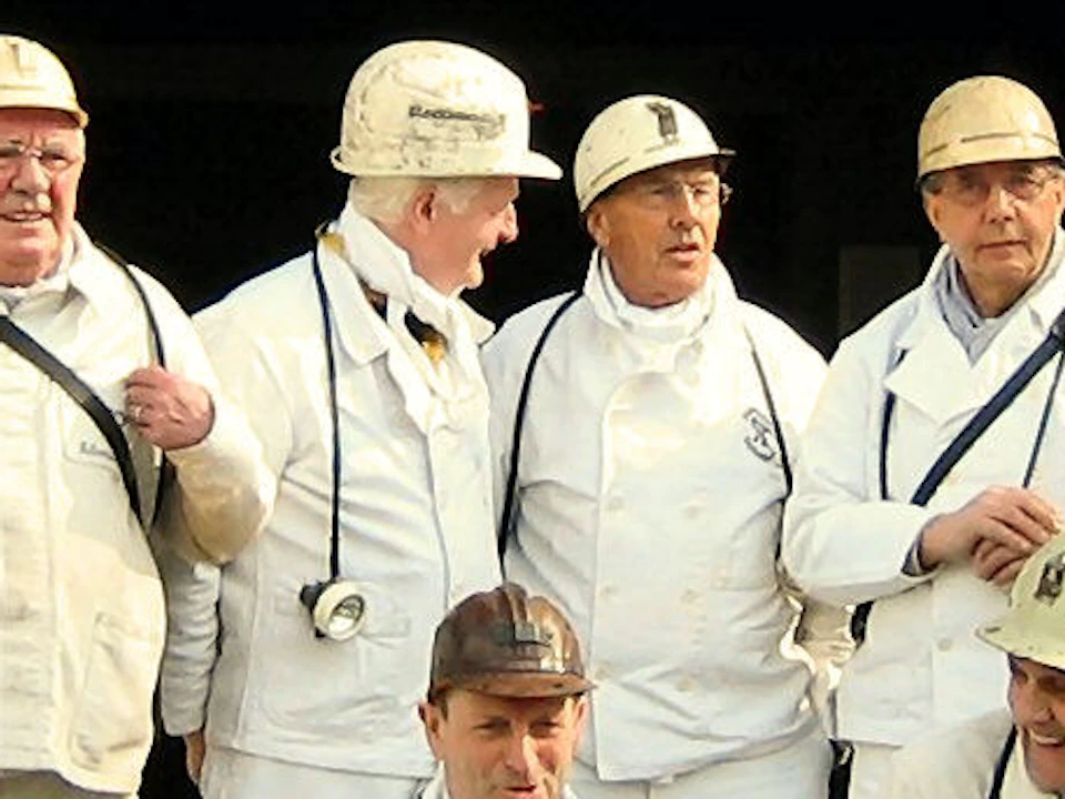 A group of people in white uniforms and helmets stand in front of a mine entrance with the sign "STEENKOLENMIJN."