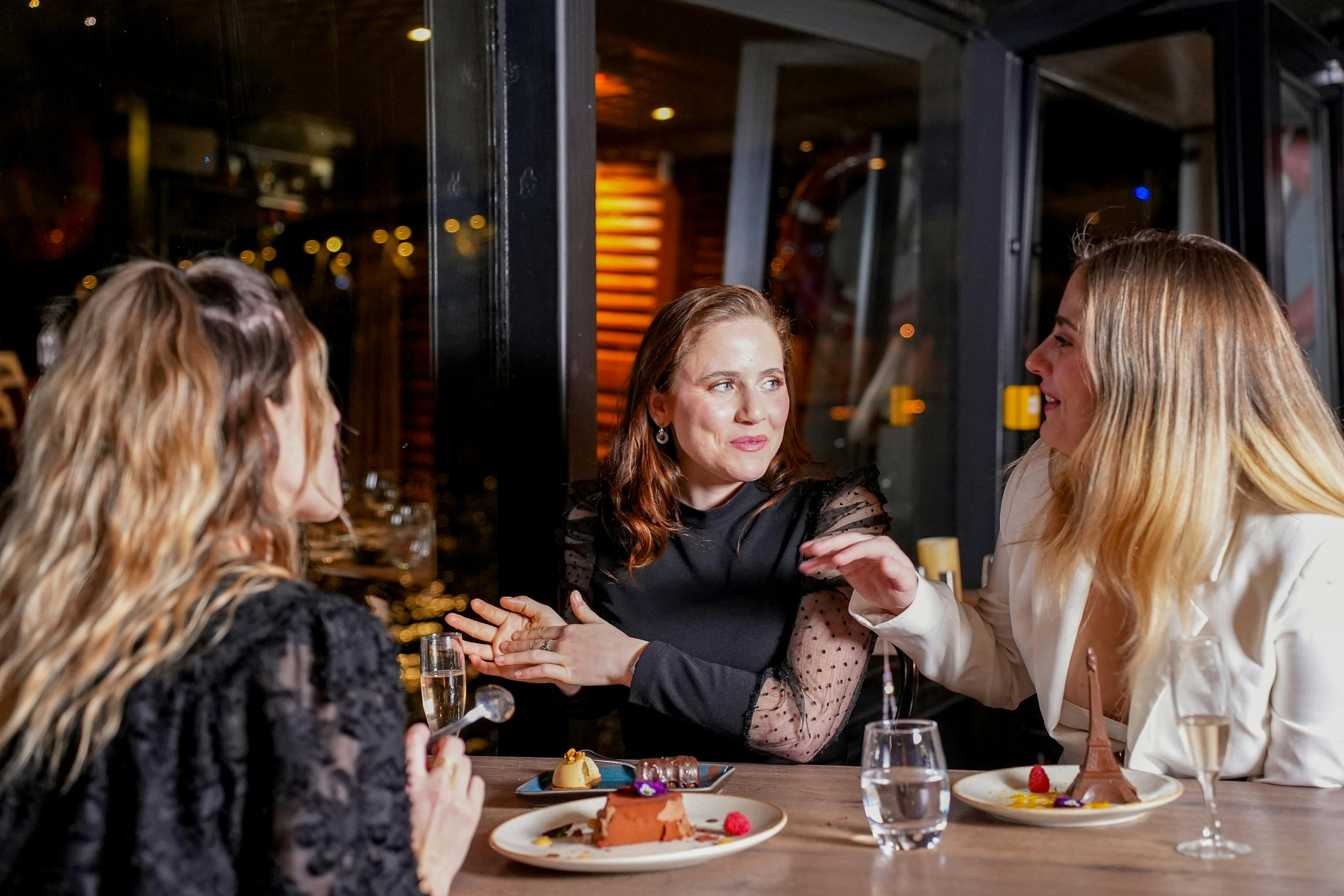 Zwei Frauen sitzen an einem Tisch in einem Restaurant, unterhalten sich und haben Dessertteller und Gläser vor sich stehen.