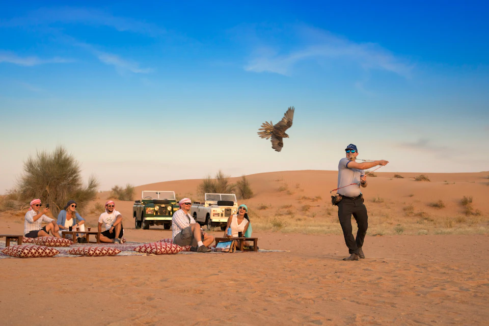 A group of people sitting in a desert with two off-road vehicles in the background and a person handling a flying bird.