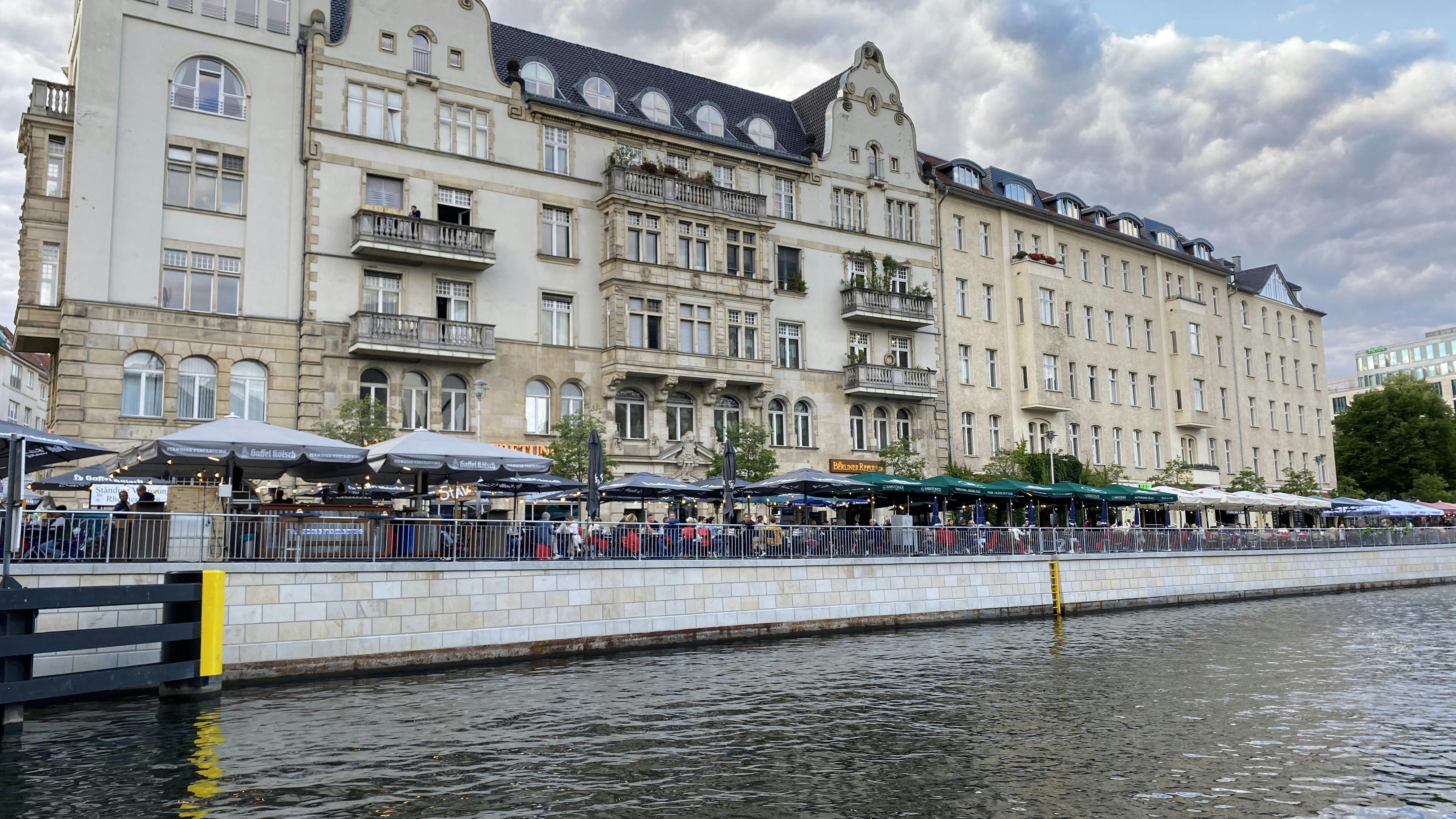 Κτίρια ιστορικού ευρωπαϊκού στιλ πλαισιώνουν μια όχθη ποταμού με υπαίθριες καφετέριες κάτω από ομπρέλες και οι άνθρωποι δειπνούν.