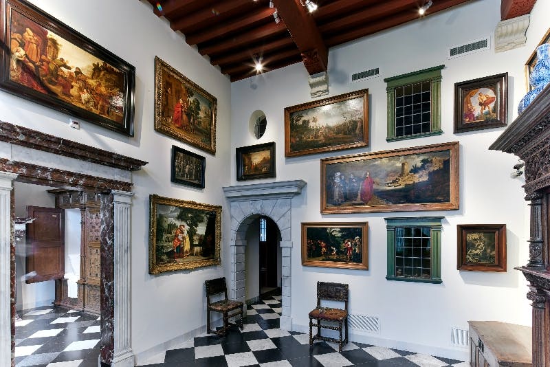 Ein Galerieraum mit Gemälden an den Wänden, einer gewölbten Tür und Stühlen auf einem karierten Boden unter einer Holzdecke.