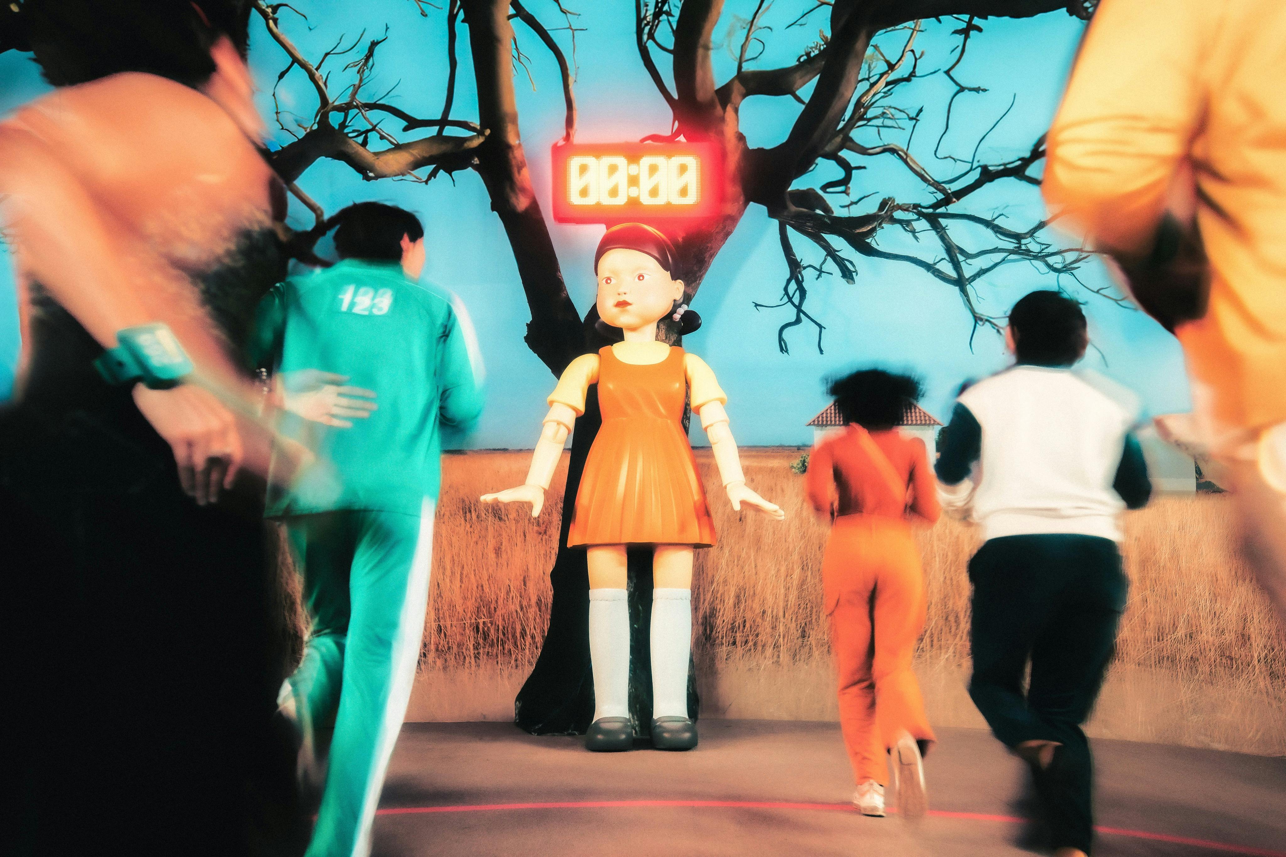 People in colorful tracksuits running towards a large doll with a timer showing 00:00 against a backdrop of leafless trees.