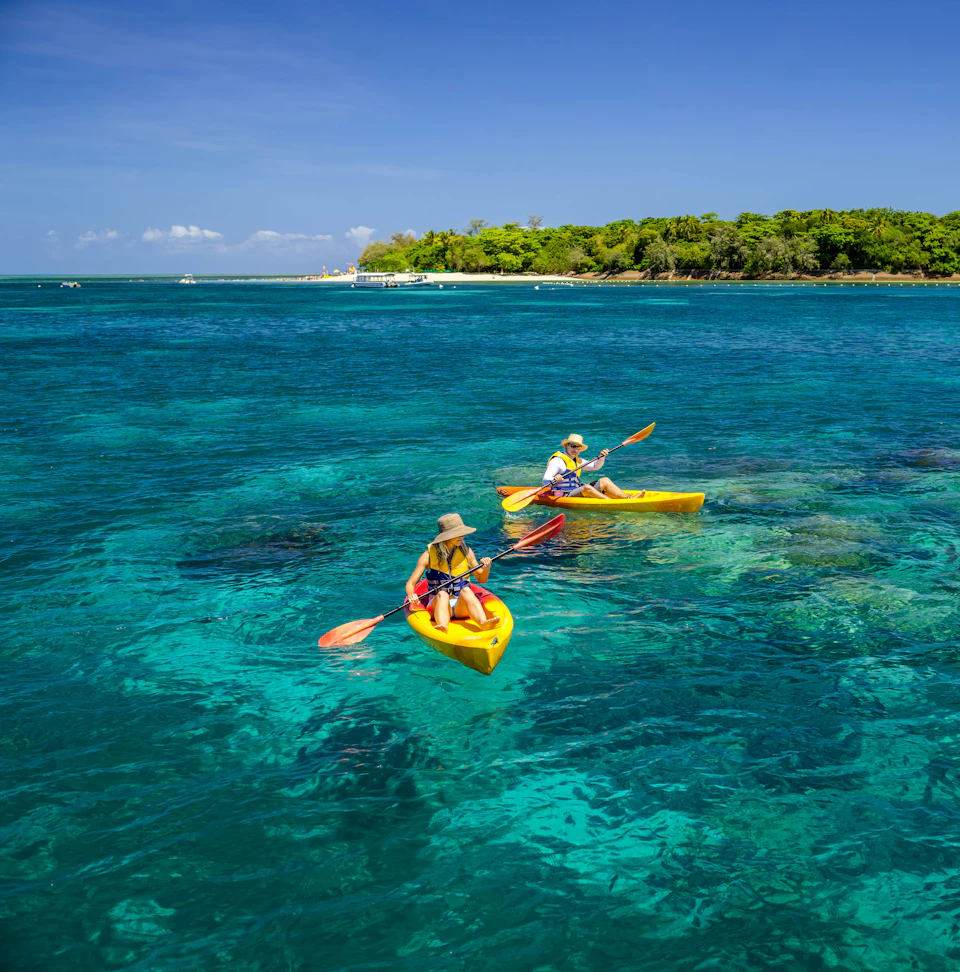 Two people in yellow kayaks paddling on clear, blue-green water near a green, tree-covered island under a bright blue sky.