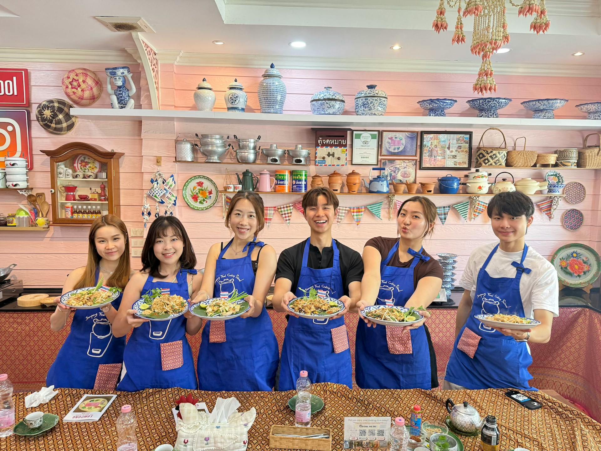 Five people wearing blue aprons stand in a kitchen holding plates of food, with colorful decor and shelves behind them.