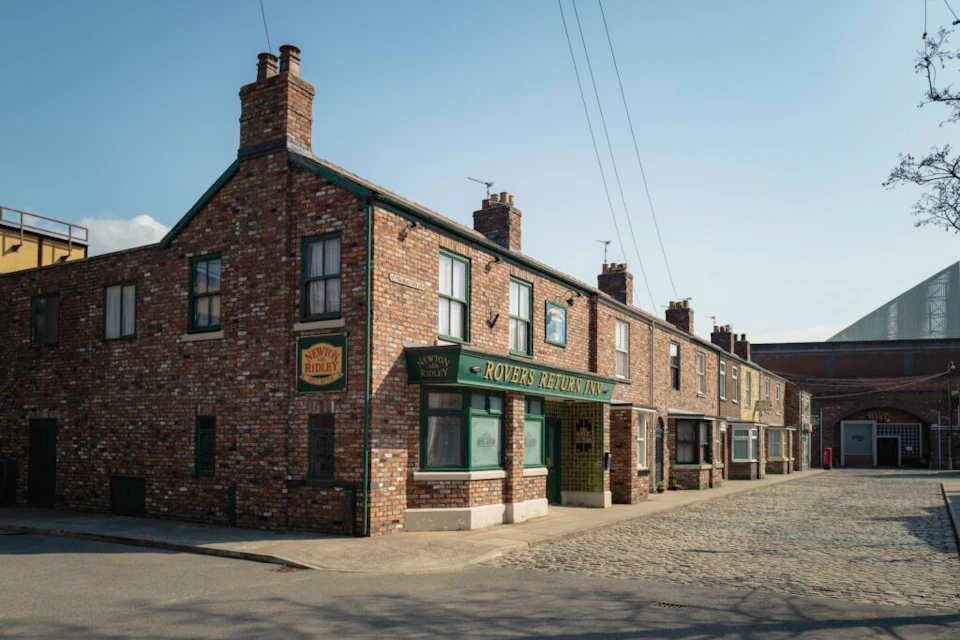 Street view of a row of brick buildings featuring the "Rovers Return Inn" pub on a sunny day. Cobblestone street in the foreground.