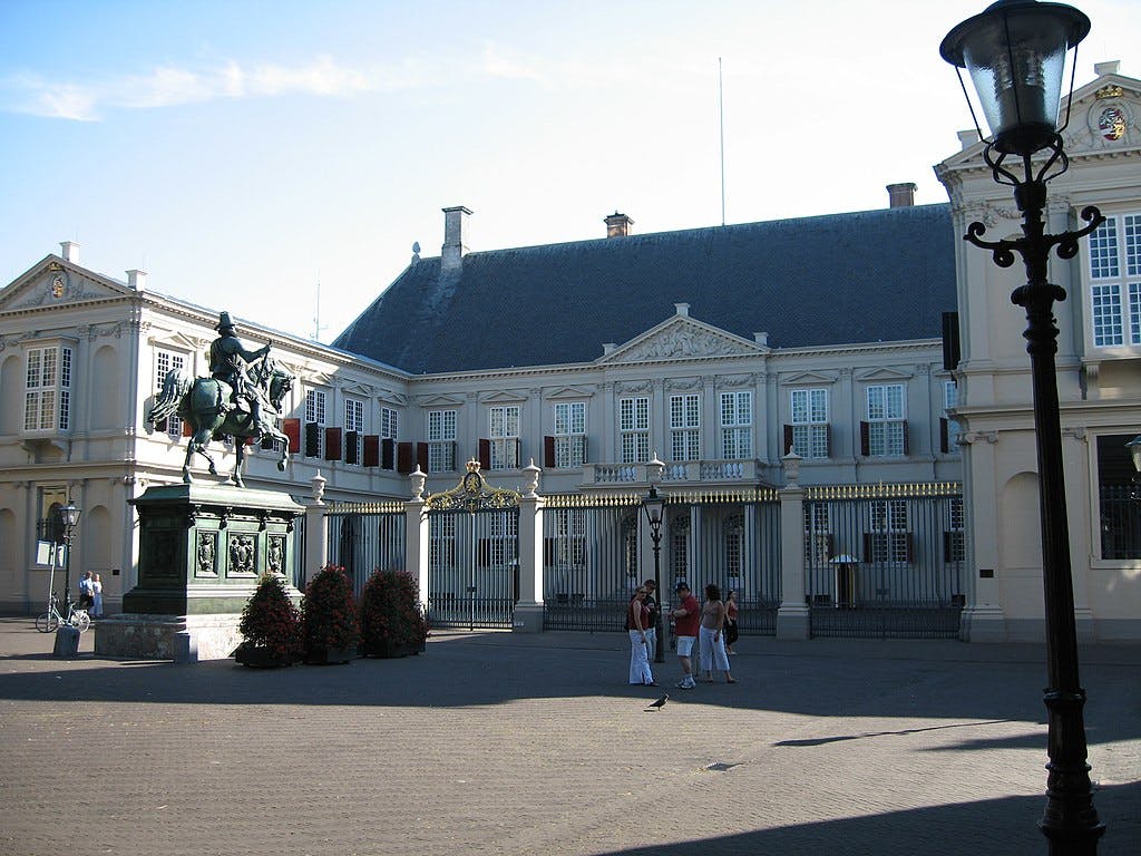 Historic building with a statue of a rider on a horse in front, people standing nearby, and a traditional lamp post.