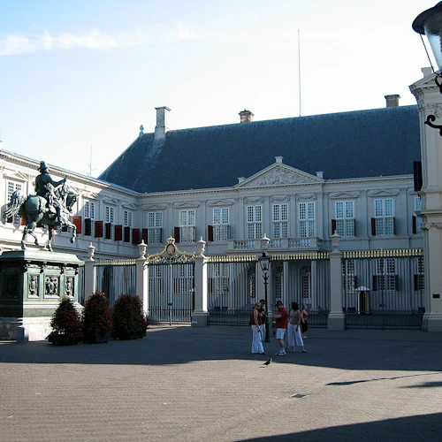 Historisk byggnad med en staty av en ryttare på en häst framför, människor som står i närheten och en traditionell lyktstolpe.