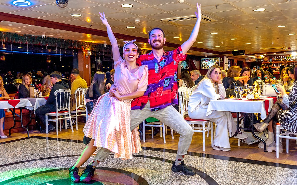 Two performers dancing in a restaurant with seated patrons watching. The space is well-lit with a lively atmosphere.