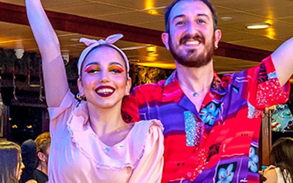 Two performers dancing in a restaurant with seated patrons watching. The space is well-lit with a lively atmosphere.