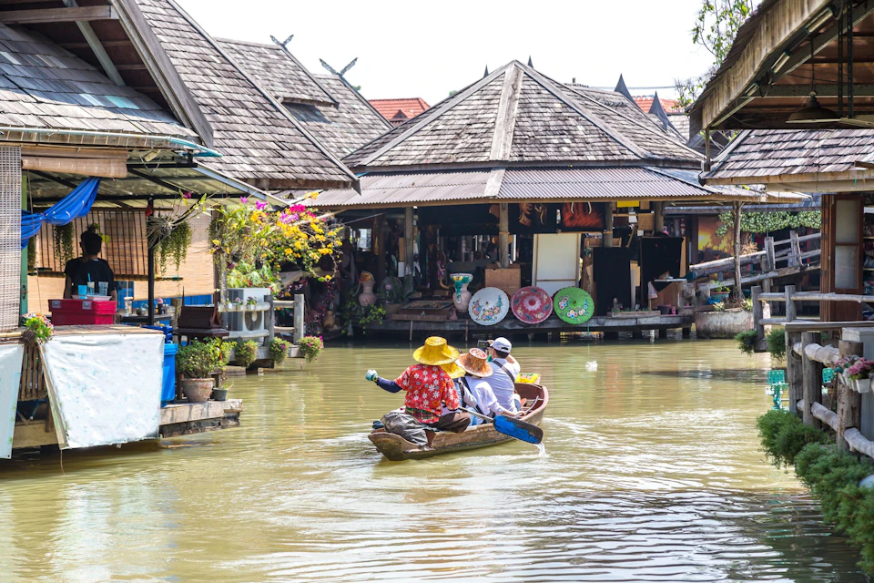 People in a small wooden boat on a canal in front of a floating market with traditional wooden buildings and goods on display.