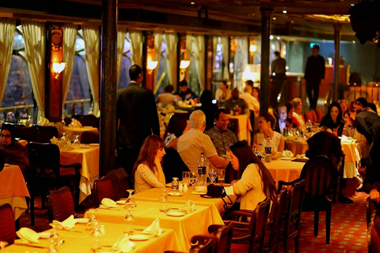 Hřejivě osvětlená, přeplněná restaurace s lidmi sedícími u stolů, kteří se zabývají konverzací a obsluhou, která podává jídlo a nápoje.