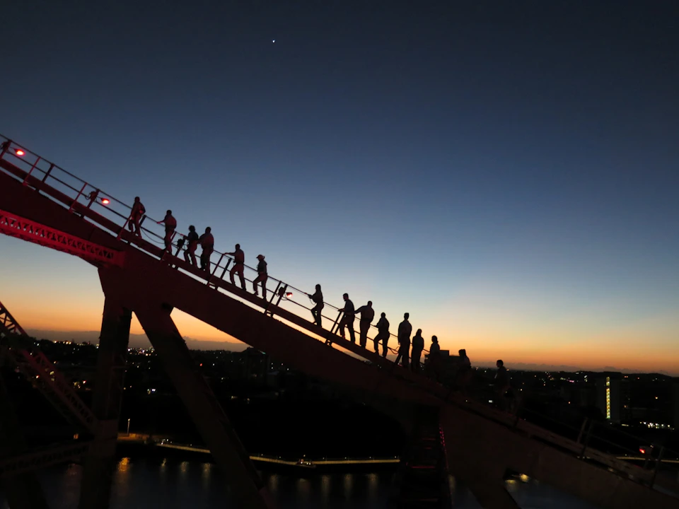 Silhouetted people climb a bridge at dusk with city lights and a deep blue sky in the background.